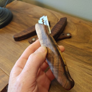 May include: A wooden razor handle with a metal razor blade inserted. The handle is made of dark wood with a light brown stripe running down the center. The razor is held in a hand.