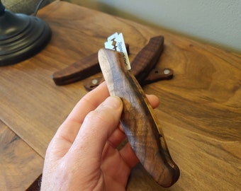 May include: A wooden razor handle with a metal razor blade inserted. The handle is made of dark wood with a light brown stripe running down the center. The razor is held in a hand.