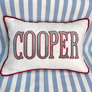 May include: A rectangular white throw pillow featuring the name COOPER embroidered in large grey letters with a red outline. The pillow has a red piped border around the entire edge, creating a distinct frame for the central text.