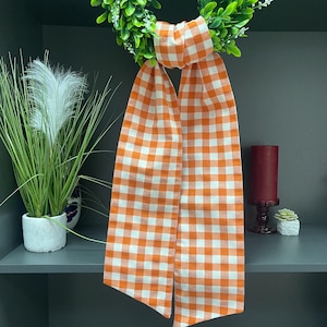 May include: A green wreath with white accents is adorned with a large orange and white gingham ribbon. The ribbon hangs down, creating a decorative element. A tall, decorative plant and a burgundy candle are also visible.