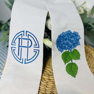May include: White graduation stole with blue embroidered details. One side features a monogram in a circular design, the other a blue hydrangea flower with green leaves. The stole is draped over a woven basket.
