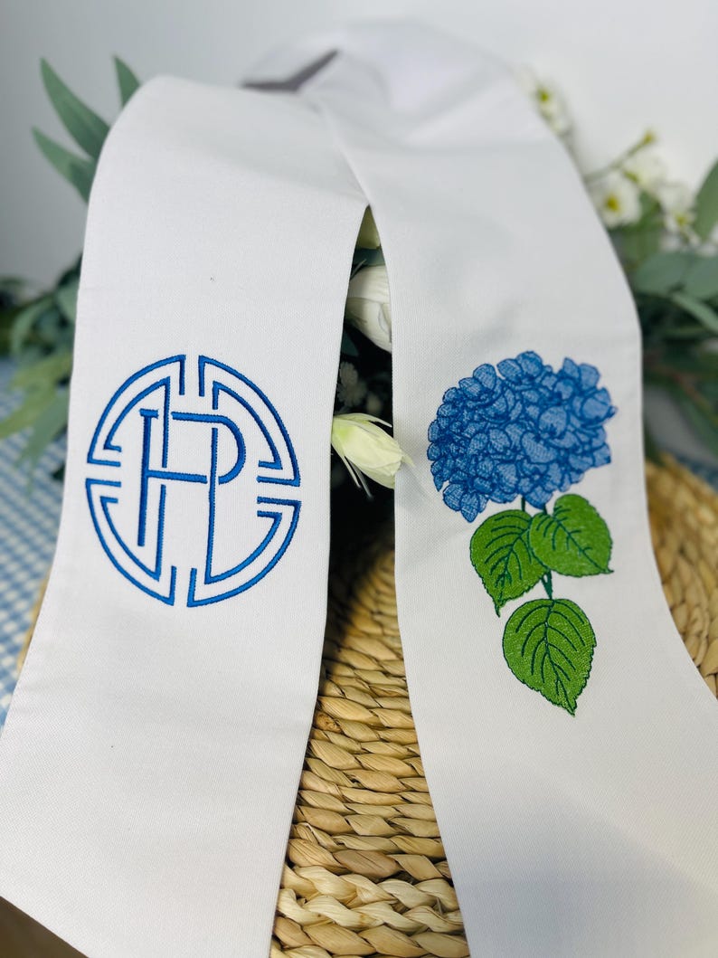 May include: White graduation stole with blue embroidered details. One side features a monogram in a circular design, the other a blue hydrangea flower with green leaves. The stole is draped over a woven basket.