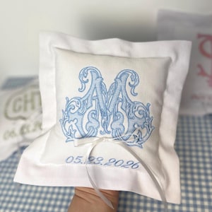 May include: A white ring pillow with a light blue embroidered monogram "M" and the date "05.23.2026". The pillow has a white ribbon and a white border. The pillow is on a blue and white checkered surface.