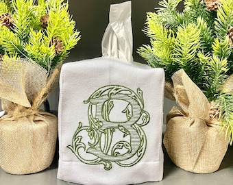 Könnte beinhalten: Weiße Taschentuchbox-Hülle mit einem gestickten grünen Buchstaben "B" in einem verzierten, floralen Design. Zwei kleine dekorative immergrüne Bäume in mit Jute umwickelten Töpfen flankieren die Box und bilden ein koordiniertes Arrangement.