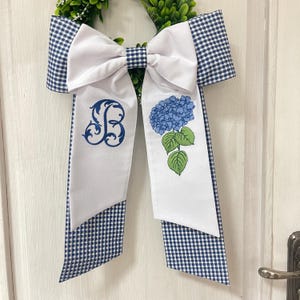 May include: A decorative wreath bow featuring a large white bow with navy blue gingham edges. The bow has two white streamers, one embroidered with a blue floral monogram and the other with a blue hydrangea design. The wreath is adorned with green foliage.