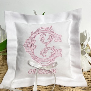 Peut inclure: Un coussin d'alliances blanc avec une lettre "C" rose brodée dans une écriture ornée. Le coussin a un ruban blanc et la date "09.22.2026" brodée en dessous. Le coussin est carré avec une bordure blanche.