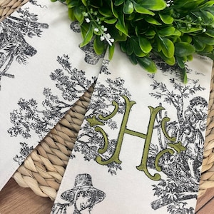 Puede incluir: Dos servilletas de tela blanquecina con un diseño de toile pastoral en blanco y negro. Una servilleta presenta una letra "H" bordada en verde. Las servilletas están sobre una superficie de mimbre tejido con follaje verde.