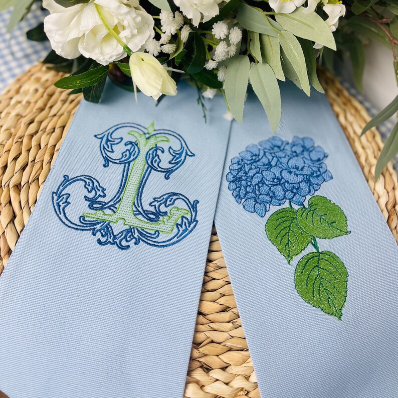 May include: Two light blue decorative towels. One towel features an embroidered letter "L" in green and blue. The other towel has a blue hydrangea flower with green leaves. The towels are displayed on a woven surface with white flowers.
