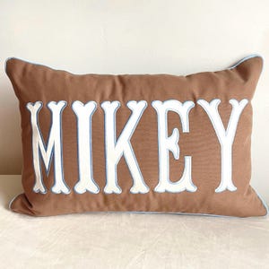 May include: A rectangular brown pillow with the name "MIKEY" embroidered in large white letters with a light blue outline. The pillow has a light blue trim around the edges.