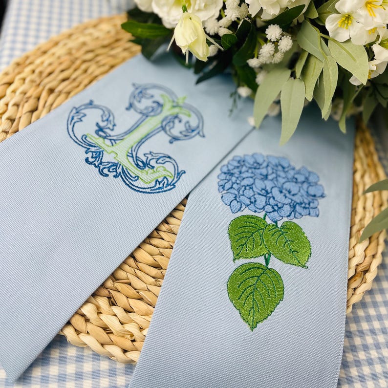 May include: Two light blue fabric ribbons with embroidered designs. One features a floral initial "J" in green and blue, the other a blue hydrangea flower with green leaves. The ribbons are displayed on a woven mat with white flowers.
