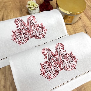 Puede incluir: Dos toallas de mano de lino blanco con una inicial "M" bordada en rojo. Las toallas tienen un borde festoneado. Una vela y una pequeña suculenta son visibles en el fondo.