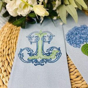 May include: Light blue linen napkins with embroidered designs. One napkin features a stylized letter "J" in blue and green, while the other has a blue hydrangea flower. The napkins are placed on a woven surface with white flowers in the background.