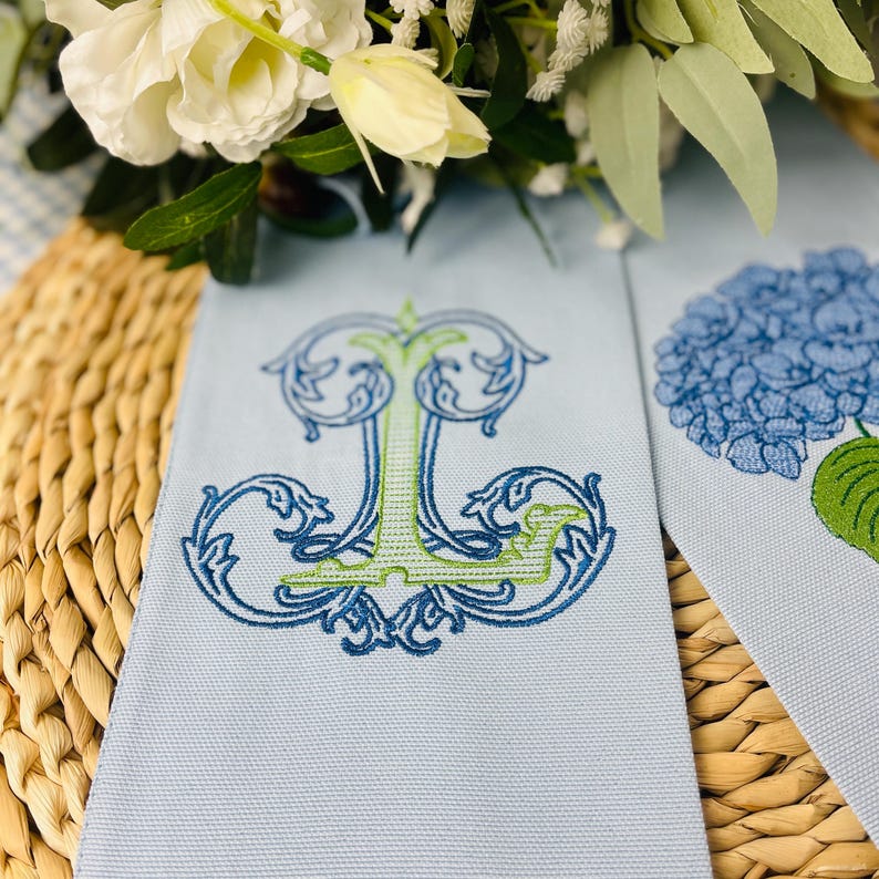 May include: Light blue linen napkins with embroidered designs. One napkin features a stylized letter "J" in blue and green, while the other has a blue hydrangea flower. The napkins are placed on a woven surface with white flowers in the background.