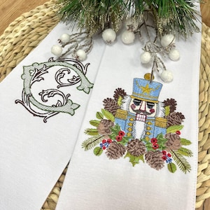 Puede incluir: Dos toallas decorativas blancas con diseños bordados. Una presenta una letra "C" verde y marrón, la otra un cascanueces azul y dorado rodeado de piñas, bayas y vegetación. Decoración festiva navideña.