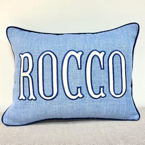 May include: A rectangular light blue throw cushion with a textured fabric finish and dark blue piping along the edges. The name ROCCO is embroidered in large, white, block-style capital letters with a dark blue outline centred on the front.
