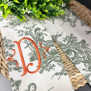 以下が含まれることがあります： クリーム色の生地に、緑色の牧歌的なデザインとオレンジ色の刺繍文字「D」が施されています。生地は、人物、木、蝶を描いたトワル柄で装飾されています。生地は編まれたバスケットの上に置かれています。