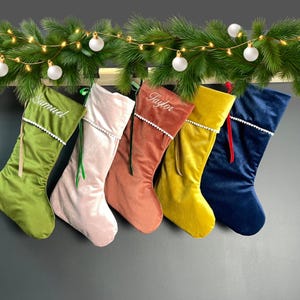 Puede incluir: Seis calcetines navideños de terciopelo en varios colores, incluyendo verde, rosa y amarillo. Cada calcetín tiene un ribete blanco y un nombre bordado. Los calcetines cuelgan de una viga de madera adornada con vegetación y adornos blancos.