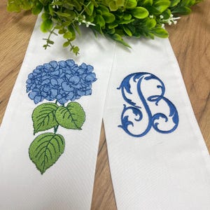 May include: Two white satin ribbons with embroidered designs. One ribbon features a blue hydrangea flower with green leaves. The other has a blue, ornate letter "B". Green foliage is in the background.