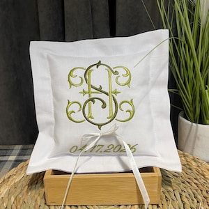 May include: White square ring pillow with an embroidered monogram in olive green and gold. The pillow has a decorative border and a satin ribbon. The date 04.17.2026 is embroidered below the monogram.