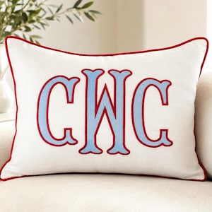 Puede incluir: Un cojín rectangular de color blanco roto que presenta un monograma bordado centrado con las letras CWC en azul claro. Las letras están delineadas con un hilo rojo intenso y todo el cojín está rematado con un ribete rojo a juego.