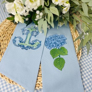 May include: Two light blue decorative towels. One towel features an embroidered letter "T" in green and blue. The other towel has a blue hydrangea flower with green leaves. The towels are on a woven mat with flowers in the background.