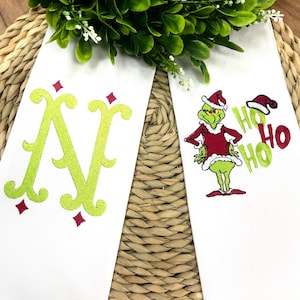 Puede incluir: Dos toallas de mano blancas con temática navideña. Una toalla presenta una gran letra "N" verde con detalles en rojo. La otra toalla tiene un personaje bordado del Grinch con un traje de Papá Noel con el texto "HO HO HO".