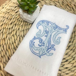 Puede incluir: Toalla de mano de lino blanco con una gran letra "T" bordada en tonos azules. La toalla está doblada y colocada sobre una estera tejida de color natural, con una pequeña planta suculenta en maceta en el fondo.