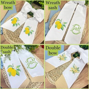 May include: Four white wreath bows and sashes with embroidered designs. Two bows feature a lemon branch design, while the other two sashes have a lemon branch and a green monogram. The bows and sashes are displayed on a woven surface.