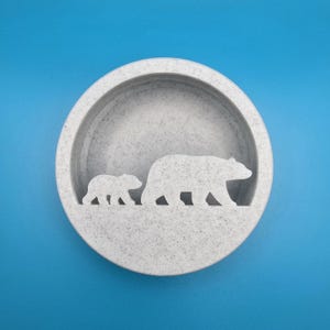 May include: A circular, pale grey decorative bowl featuring a bear and cub silhouette. The bowl has a textured finish and is set against a bright blue backdrop.