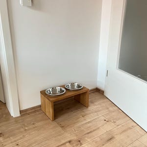 Puede incluir: Una estaci&oacute;n de alimentaci&oacute;n para mascotas de madera con dos cuencos de acero inoxidable. Los cuencos est&aacute;n sobre un peque&ntilde;o soporte rectangular de madera. El soporte est&aacute; sobre un suelo de parquet. Una pared blanca y un marco de puerta est&aacute;n en el fondo.