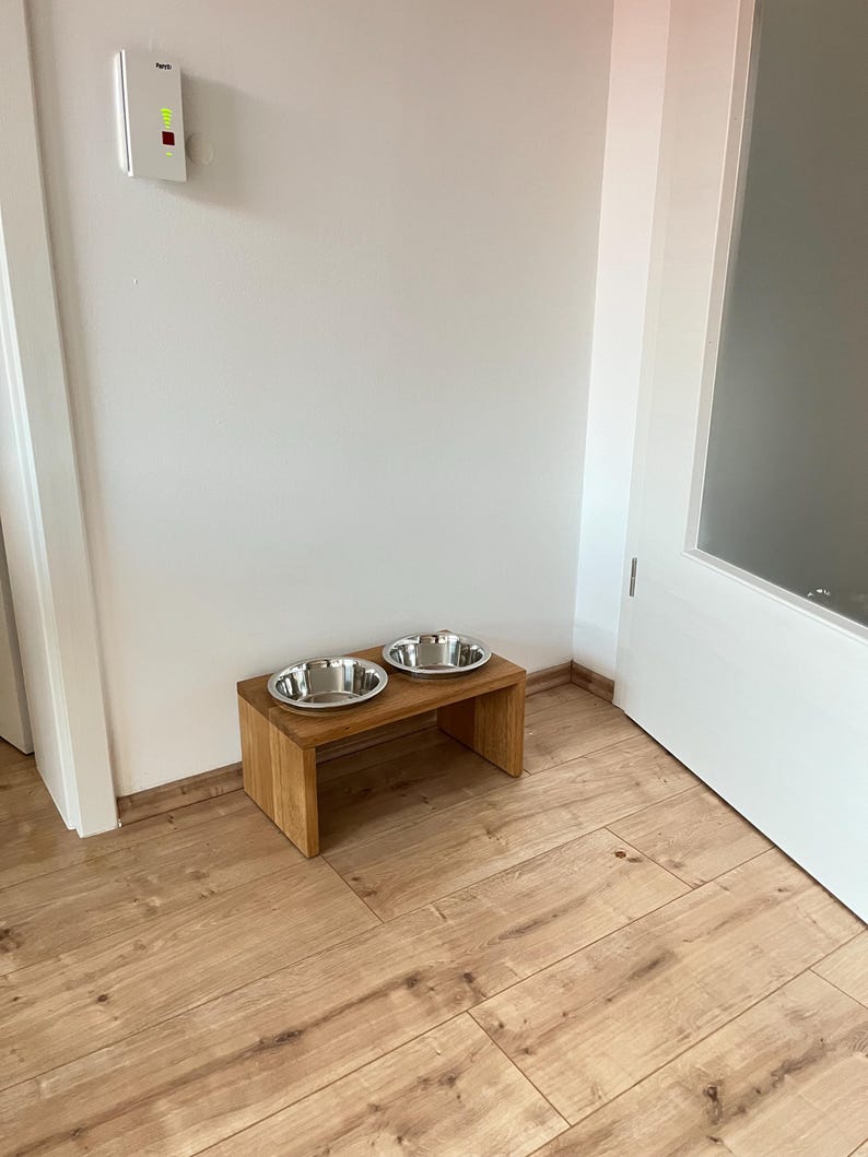 Puede incluir: Una estaci&oacute;n de alimentaci&oacute;n para mascotas de madera con dos cuencos de acero inoxidable. Los cuencos est&aacute;n sobre un peque&ntilde;o soporte rectangular de madera. El soporte est&aacute; sobre un suelo de parquet. Una pared blanca y un marco de puerta est&aacute;n en el fondo.