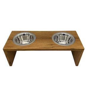Puede incluir: Un comedero de madera para mascotas con dos cuencos de acero inoxidable. El comedero est&aacute; hecho de madera de color marr&oacute;n claro y tiene una parte superior rectangular con dos recortes circulares para los cuencos. Los cuencos son brillantes y reflejan la luz. El comedero est&aacute; elevado con patas anguladas.