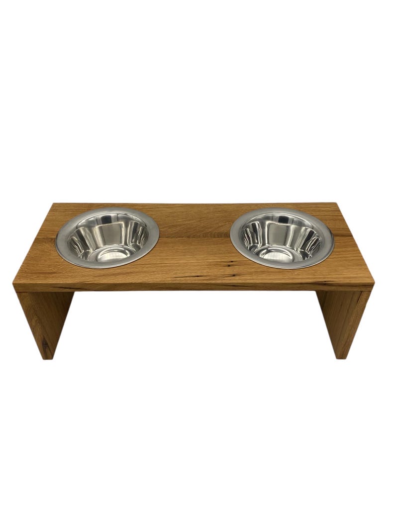 Puede incluir: Un comedero de madera para mascotas con dos cuencos de acero inoxidable. El comedero est&aacute; hecho de madera de color marr&oacute;n claro y tiene una parte superior rectangular con dos recortes circulares para los cuencos. Los cuencos son brillantes y reflejan la luz. El comedero est&aacute; elevado con patas anguladas.