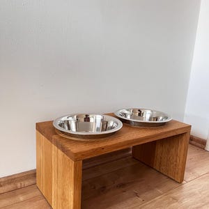 Puede incluir: Un comedero de madera para mascotas con dos cuencos de acero inoxidable. El comedero es de madera marr&oacute;n claro y tiene un dise&ntilde;o simple y moderno. Los cuencos son redondos y brillantes, ideales para alimentar a las mascotas.