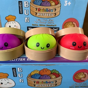 May include: Three squishy bun toys in individual tan plastic steamer baskets. The buns are purple, lime green, and red, each featuring a printed smiling face with pink cheeks. They are displayed on a blue cardboard box labeled Squishy Bun.