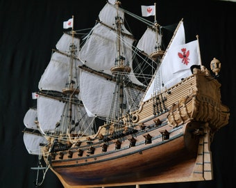 May include: A detailed wooden model of a sailing ship with white sails and red flags. The ship has intricate carvings and details, including cannons and rigging. The model is displayed on a black background.
