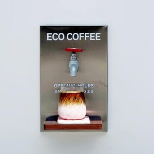 Op de afbeelding: Een roestvrijstalen koffieautomaat met de tekst "ECO COFFEE" boven een kraan en een glas ijskoffie. Het glas staat op een klein houten plankje met een roze onderzetter. De openingstijden worden weergegeven.