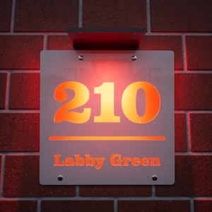 以下が含まれることがあります： オレンジ色の数字210と「Labby Green」という名前が書かれた、照明付きの家の番号看板。 赤レンガの壁に取り付けられ、温かいオレンジ色の光で照らされています。