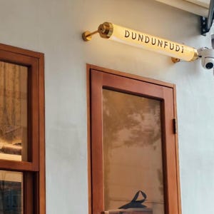 Könnte beinhalten: Außenansicht eines Gebäudes mit Holztür und einem Schild mit der Aufschrift "DUNDUNFUDI". Das Schild ist eine zylindrische Leuchte mit goldenen Akzenten. Rechts vom Schild ist eine Überwachungskamera angebracht.