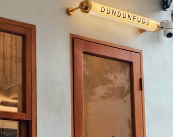 May include: Exterior shot of a building with a wooden door and a sign that reads "DUNDUNFUDI". The sign is a cylindrical light fixture with gold accents. A security camera is mounted to the right of the sign.