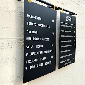 May include: Two black rectangular menu boards with white lettering hanging from a black metal bar. The left board lists items like Margherita, Tomato Mozzarella, Calzone, Mushroom & Cheese, Spicy Nduja, and Hazelnut Pesto with prices.