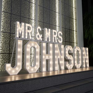 May include: A large illuminated sign reading "MR & MRS JOHNSON" in white marquee letters. The sign, lit with bulbs, is likely for a wedding. The background features a decorative wall.