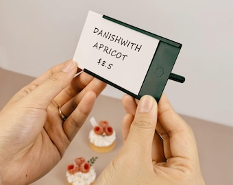 Puede incluir: Un soporte de metal verde oscuro que muestra una tarjeta rectangular blanca con el texto impreso en negro DANISH WITH APRICOT y $8.5. El soporte presenta un pequeño logotipo hexagonal y una varilla horizontal en el lado derecho.
