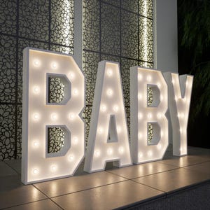 May include: Large white marquee letters spelling "BABY" with warm white lights. The letters are illuminated and stand on a tiled surface, with a decorative patterned wall in the background. A festive decoration for a baby shower or nursery.