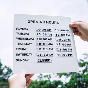 May include: A white square sign with black text reading OPENING HOURS, listing Monday through Sunday with specific times. Monday through Friday show 10:00 AM to 12:00 PM, Saturday shows 11:30 AM to 20:00 PM, and Sunday is marked as CLOSED-.