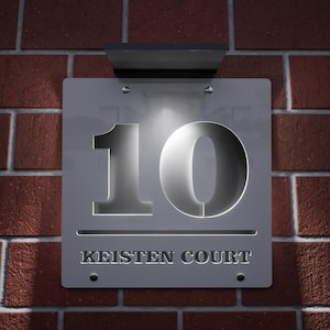 以下が含まれることがあります： 赤いレンガの壁に取り付けられた、数字の10と「KEISTEN COURT」の文字が入った銀色の金属製住所プレート。 プレートの上には小さな照明器具があります。