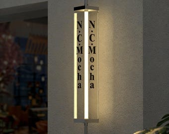 May include: A rectangular, illuminated sign with the text "N C M Mocha" in black lettering. The sign is encased in a clear, vertical tube with a bright, vertical light strip. The sign is mounted on a wall with a chrome-colored, curved bracket.