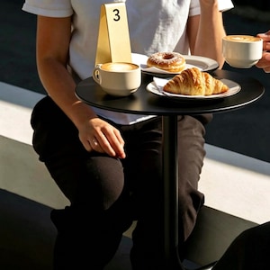 Könnte beinhalten: Ein kleiner schwarzer Tisch mit Cappuccino, Croissant und Donut. Eine goldene Tischnummer 3 befindet sich auf dem Tisch. Der Tisch befindet sich im Freien, mit einer Person, die am Tisch sitzt.