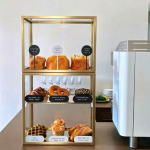 Könnte beinhalten: Eine goldfarbene, zweistöckige Glasvitrine mit verschiedenen Backwaren auf den Regalen. Die Vitrine ist mit kleinen runden und ovalen Schildern versehen, die Textbeschriftungen für Artikel wie Croissant, Karottenkuchen und Waffel enthalten.