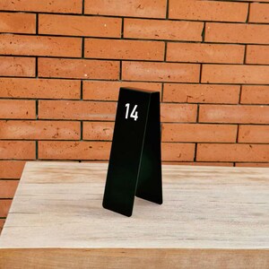 May include: A black A-frame table number sign featuring the white number 14 printed on the front panel. The sign has a smooth, matte finish and a triangular profile, designed to stand upright on a flat surface for clear visibility in settings.
